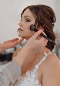 Eine Frau mit hochgestecktem braunem Haar lässt sich mit einem Pinsel Make-up auf die Wangen auftragen und bereitet sich auf ihre Hochzeit vor. Sie trägt ein weißes Spitzenkleid mit dünnen Trägern, perfekt für diesen Anlass. Der Hintergrund ist verschwommen und lässt auf eine Innenumgebung schließen, in der sorgfältige Hochzeitsplanung stattfindet.