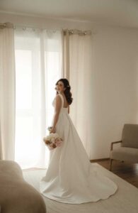 Eine Braut in einem fließenden weißen Kleid steht vor transparenten Vorhängen und hält einen Strauß rosa und weißer Blumen in der Hand. Der Raum, der sich perfekt für die Hochzeitsplanung eignet, ist in sanften, neutralen Tönen gehalten und bietet mit einem flauschigen Teppich und einem Sessel im Hintergrund die perfekte Kulisse für eine unvergessliche Hochzeit.