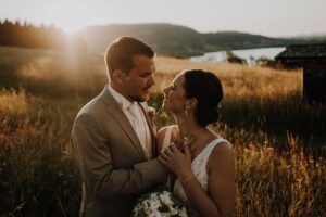 Ein Paar in Abendgarderobe steht auf einem sonnenbeschienenen Feld, perfekt für die Hochzeitsplanung, mit sanften Hügeln und einem schimmernden Gewässer im Hintergrund. Der Bräutigam hält die Hände der Braut, während sie sich liebevoll anschauen. Sonnenlicht fällt durch das hohe Gras und fängt ihren magischen Hochzeitstag ein.