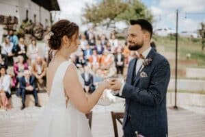 Während ihrer bezaubernden Hochzeit tauschen Braut und Bräutigam unter freiem Himmel die Ringe aus. Sie stehen einander gegenüber, umgeben von Gästen und einer atemberaubenden Kulisse aus Bäumen. Die Braut trägt ein makelloses weißes Kleid, während der Bräutigam einen schicken blauen Anzug trägt, was eine idyllische Szene für die Hochzeit schafft.
