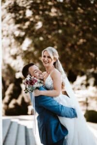 Ein Bräutigam im blauen Anzug hebt freudig die lächelnde Braut hoch, die ein weißes Brautkleid trägt und einen Blumenstrauß hält. Sie feiern ihre Hochzeit im Freien inmitten verschwommenen Grüns und sonnen sich an ihrem besonderen Tag.