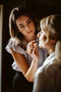 Zur Vorbereitung einer Hochzeit trägt eine Visagistin einer sitzenden Frau Lippenstift auf. Die Visagistin ist konzentriert und trägt ein leichtes, gerüschtes Oberteil. Die sanft beleuchtete Umgebung schafft eine intime Atmosphäre, perfekt für die Heiratsstimmung des Tages.