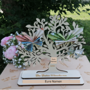 Eine dekorative Baumskulptur mit Papierschmetterlingen auf einem Holzsockel schmückt den Tisch, umgeben von rosa Rosen und üppigem Grün. Mit den Aufschriften „Die besten Wünsche von“ und „Eure Namen“ ist sie ein perfektes Stück für Ihre Hochzeitsplanung.