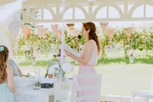 Eine Frau in einem rosa Kleid steht mit Papieren an einem Mikrofon während einer Hochzeit im Freien, die mit dekorativen Fahnen und Blumen geschmückt ist. Die Umgebung wirkt hell und sonnig, mit viel Grün und Weinreben im Hintergrund. Die Gäste sitzen an einem wunderschön dekorierten Tisch.