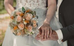 Eine Braut mit einem Strauß blassrosa Rosen und Grünzeug steht neben einem Bräutigam im schwarzen Anzug, der sanft seine Hand auf ihre legt. Beide zeigen stolz ihre Eheringe, die ihre Verbindung symbolisieren, die mit sorgfältiger Hochzeitsplanung begann.