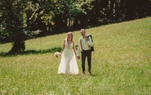 Eine Braut im weißen Kleid und ein Bräutigam mit Hosenträgern und einer Jacke über der Schulter gehen Hand in Hand über eine Wiese und verkörpern damit die Essenz einer Hochzeit. Sie hält einen Blumenstrauß und beide lächeln unter einem sonnigen Himmel mit Bäumen im Hintergrund.