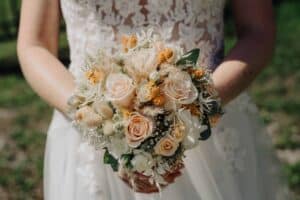 Eine Braut, die einen Strauß pfirsichfarbener und cremefarbener Rosen, gemischt mit weißen und gelben Blumen und Grünzeug, in der Hand hält, verkörpert die Essenz einer Traumhochzeit. Sie trägt ein weißes Kleid mit Spitzendetails, das Eleganz ausstrahlt. Der Hintergrund ist sanft verschwommen und verleiht einen Hauch von romantischem Mysterium.