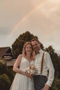 Eine Braut in einem weißen Blumenkleid und ein Bräutigam in einem weißen Hemd mit Hosenträgern stehen lächelnd da und halten einen Blumenstrauß in der Hand. Sie sind draußen, mit einem Haus und Bäumen im Hintergrund, unter einem Himmel, der einen schwachen Regenbogen zeigt – ein bildschöner Moment ihrer Hochzeit.