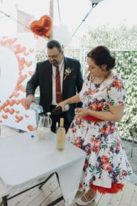 Ein Mann im Anzug und eine Frau im geblümten Kleid stehen an einem elegant gedeckten Tisch im Freien. Als Teil ihrer Hochzeitsplanung gießen sie Flüssigkeiten aus Flaschen in einen Krug, umgeben von herzförmigen Luftballons und Herzdekorationen, und schaffen so die perfekte Bühne für ihre Hochzeit.