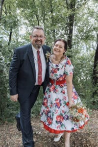 Ein Paar steht an seinem Hochzeitstag im Freien und lächelt breit. Der Mann trägt einen dunklen Anzug mit roter Krawatte und die Frau ein geblümtes Kleid mit roten Akzenten und hält einen Blumenstrauß in der Hand. Bäume und Grün umgeben sie und bilden eine perfekte Kulisse für ihre wunderschöne Hochzeitsfeier.