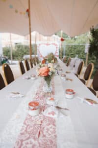 Ein langer, eleganter Tisch ist für eine formelle Hochzeit unter einem großen Zelt gedeckt. Er verfügt über einen Tischläufer aus Spitze, ein Blumengesteck mit Rosen, kleine dekorierte Gefäße und Besteck. Im Hintergrund ist ein Hintergrund mit einem großen Herz zu sehen, der diesem Hochzeits-Ambiente Charme verleiht.