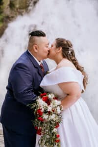 Ein Paar in Abendgarderobe küsst sich vor einem Wasserfall und feiert seine Hochzeit. Der Bräutigam trägt einen marineblauen Anzug und die Braut glänzt in einem weißen schulterfreien Kleid und hält einen Strauß roter und weißer Blumen in der Hand.