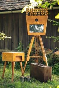Eine charmante Vintage-Fotokabine im Freien, perfekt für Ihre Hochzeit, mit einer Holzkamera auf einem Stativ, einem Lautsprecher im Retro-Stil auf einem Ständer und einem braunen Koffer. Das Wort „Fotos“ ist oben vor dem Hintergrund des rustikalen Holzgebäudes deutlich zu sehen.