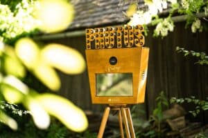Eine Fotokabine im Vintage-Stil, perfekt für die Hochzeitsplanung, hat eine Holzfassade und steht im Freien auf einem Stativ. Geschmückt mit Laufschriftbuchstaben mit der Aufschrift „PHOTOS“, fängt sie das durch die Blätter fallende Sonnenlicht ein und schafft eine warme Atmosphäre, die sich ideal für jede Hochzeitsumgebung eignet.