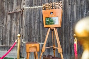 Eine Fotokabine im Vintage-Stil mit einem Holzstativ und einem beleuchteten „Fotos“-Schild wird im Freien aufgestellt und verleiht jeder Hochzeit Charme. Sie steht neben einem roten Samtseil und goldenen Pfosten vor einer rustikalen Holzwand. Daneben steht eine kleine Box mit einer Schublade, perfekt zum Festhalten von Hochzeitsmomenten.