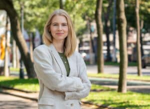 Eine Frau mit blondem Haar steht selbstbewusst da, die Arme verschränkt, in einem hellen Blazer und einem grünen Oberteil. Vor einer parkähnlichen Kulisse, in der Bäume und Straßen hinter ihr sanft verschwimmen, strahlt sie die ruhige Sicherheit einer Person aus, die in die Hochzeitsplanung vertieft ist.