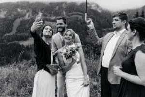 Eine Gruppe von fünf Personen, darunter eine Braut im Hochzeitskleid, posiert glücklich für ein Selfie inmitten malerischer Berge und feiert ihre Hochzeit. Eine Person hält in diesem Schwarz-Weiß-Schnappschuss einen Regenschirm.