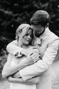 Ein Schwarzweißfoto zeigt ein Paar, das sich im Freien umarmt und die Freude einer Hochzeit verkörpert. Der Mann im Anzug hält die Frau, die ein Spitzenkleid trägt und zufrieden die Augen geschlossen hat, sanft von hinten. Üppiges Grün verschwimmt romantisch im Hintergrund.