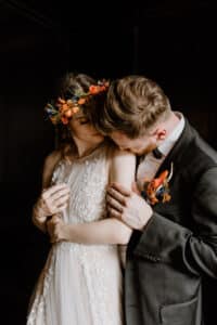 Inmitten ihrer bezaubernden Hochzeit erleben Braut und Bräutigam einen zärtlichen Moment. Die Braut, in einem weißen Blumenkleid und mit einem bunten Blumenkranz, wird von ihrem Bräutigam, der einen schwarzen Anzug mit einer Blumenboutonniere trägt, sanft auf den Arm geküsst.