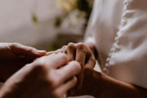 Nahaufnahme zweier Hände, von denen eine während eines intimen Heiratsmoments sanft einen Ring an den Finger der anderen steckt. Die Person, die den Ring erhält, trägt ein weißes Kleidungsstück mit Knopfdetails vor einem leicht verschwommenen Hintergrund.