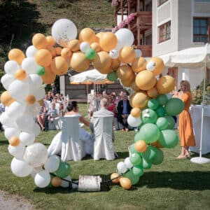 Ein bunter Ballonbogen umrahmt eine Hochzeitszeremonie im Freien. Die Gäste sitzen und eine Frau in einem orangefarbenen Kleid steht daneben. Weiße Stühle und ein Rasen vervollständigen die Szene, mit einem Gebäude im Hintergrund, das tadellose Hochzeitsplanung zur Schau stellt.