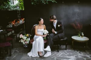 Eine Braut in einem weißen Kleid und ein Bräutigam in einem schwarzen Anzug mit Turnschuhen sitzen zusammen auf einer Bank in einer idyllischen Hochzeitsszene im Freien. Sie sind von Blumen umgeben, im Hintergrund steht ein für ihr Essen gedeckter Tisch. Die dunkle, mit Grünpflanzen geschmückte Wand verleiht der Hochzeitsszene Charme.