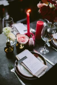 Eine romantische Tischdekoration, die perfekt für eine Hochzeit ist, besteht aus einer brennenden roten Kerze, rosa und weißen Blumen in kleinen Vasen, einer gefalteten Serviette mit einem Menü auf einem Teller und leeren Weingläsern. Die Atmosphäre ist intim mit sanfter Beleuchtung, ideal für diejenigen, die schon tief in der Hochzeitsplanung stecken.