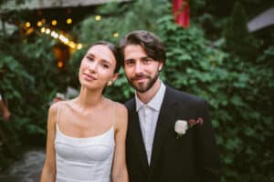 Ein Paar lächelt an seinem Hochzeitstag im Freien. Die Frau trägt ein weißes Kleid mit dünnen Trägern, während der Mann einen schwarzen Anzug mit weißem Hemd und Ansteckblume trägt. Sie stehen dicht beieinander, umgeben von Grünpflanzen und Lichterketten, und feiern die Freude des Heiratens.