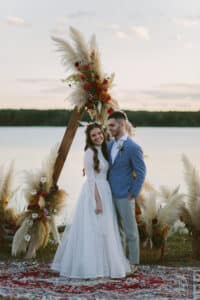 Ein Paar in Hochzeitskleidung steht vor einem dreieckigen Bogen, der mit Pampasgras und Blumen geschmückt ist, an einem Seeufer. Die Braut trägt ein weißes Spitzenkleid und der Bräutigam eine blaue Jacke. Ein gemusterter Teppich ergänzt die unkonventionelle Atmosphäre und fängt ihre Hochzeit wunderschön in dieser bezaubernden Landschaft ein.