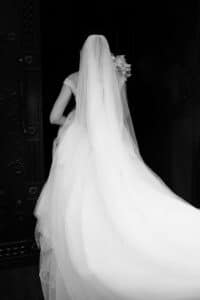 Eine Braut in einem fließenden weißen Brautkleid und Schleier steht mit dem Rücken zur Kamera. Sie hält einen Blumenstrauß in der Hand und verkörpert damit die Essenz einer Hochzeit in einem schwach beleuchteten Raum, was einen auffälligen Kontrast zu ihrem hellen Kleid bildet.