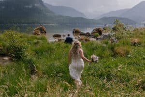 Eine Braut in einem weißen Kleid mit einem Blumenstrauß geht durch üppiges Grün zu einem ruhigen See, umgeben von Bergen unter bewölktem Himmel – eine perfekte Kulisse für eine romantische Hochzeit. In der Ferne steht jemand am Wasser und trägt zur ruhigen Landschaft bei.