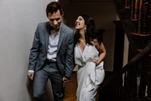 Ein lächelndes Paar geht Händchen haltend eine Treppe hinauf und strahlt die Freude über seine Hochzeit aus. Der Mann trägt einen grauen Anzug, während die Frau ein weißes Kleid trägt. Sie schauen sich glücklich an und halten einen wunderschönen Moment an ihrem besonderen Tag fest.