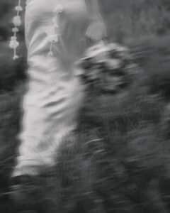 Ein unscharfes Schwarzweißbild zeigt eine Person in einem wallenden Kleid, die einen Blumenstrauß hält, und fängt die Essenz einer Hochzeit im Freien mit Gras im Vordergrund ein.