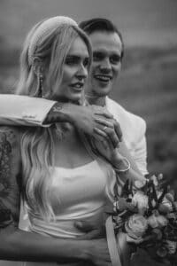 Braut und Bräutigam stehen dicht beieinander, lächeln und sind in ihre Hochzeit vertieft. Die Braut hält einen Blumenstrauß und trägt ein Perlenstirnband zu ihrem ärmellosen Kleid, während der Bräutigam in seinem Anzug sie sanft umarmt. Das Schwarzweißfoto fängt ihre Szenerie im Freien wunderschön ein.