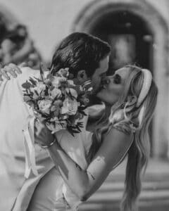 Ein Schwarzweißfoto fängt einen romantischen Moment beim Heiraten ein, während sich das Paar küsst. Die Frau mit langen Haaren und Stirnband hält einen Blumenstrauß, während der Mann sie vor einem unscharfen Außenhintergrund leicht senkt.