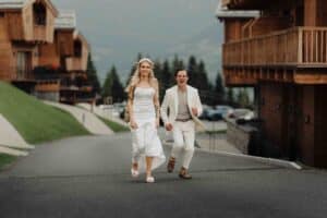 Ein Paar in weißen Outfits läuft fröhlich eine gepflasterte Straße entlang, die von Holzchalets und Grünflächen gesäumt ist, scheinbar in einem spontanen Moment inmitten ihrer Hochzeit gefangen. Der bewölkte Himmel unterstreicht die malerische Bergkulisse, während ihr langes blondes Haar weht und er ausgelassen bleibt.