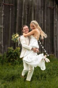 In einer malerischen Umgebung im Freien hebt ein Bräutigam seine lächelnde Braut während ihrer Hochzeit freudig hoch. Beide sind in Weiß gekleidet und sie hält anmutig einen Strauß gelber und weißer Blumen. Die Szene wird durch einen rustikalen Holzzaun im Hintergrund wunderschön ergänzt.
