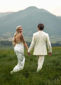 Ein Paar in eleganter weißer Kleidung geht Hand in Hand durch eine Wiese und verkörpert damit die Freude ihrer Hochzeit. Die Frau mit langen blonden Haaren und einem sichtbaren Tattoo spaziert neben ihrem Partner mit kurzen dunklen Haaren, während eine malerische Berglandschaft die perfekte Kulisse für ihren besonderen Tag bildet.