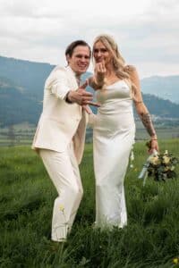 Ein Paar in Hochzeitskleidung steht auf grünem Gras mit Bergen im Hintergrund und verkörpert die Freude ihrer Hochzeit. Sie gestikulieren spielerisch in Richtung Kamera; die Braut hält einen Blumenstrauß. Beide lächeln und wirken fröhlich und fangen einen perfekten Moment ihrer Hochzeitsplanung ein.