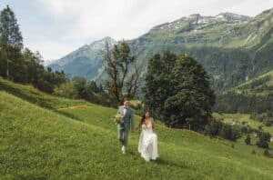 Ein Paar im Hochzeitskleid geht Hand in Hand über einen üppigen grünen Hügel und verkörpert so seine perfekte Hochzeit. Die Braut hält einen Blumenstrauß, während der Bräutigam in einem grünen Anzug hervorsticht. Majestätische Berge und Bäume bilden die malerische Kulisse unter einem teilweise bewölkten Himmel und machen diesen Moment unvergesslich.
