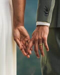 Eine Nahaufnahme von zwei Händen, die sich sanft berühren und jeweils einen goldenen Ehering tragen. Eine Person trägt ein helles Kleid und die andere einen grauen Anzug. Der sanft verschwommene Hintergrund erinnert an eine Hochzeit im Freien und fängt die Essenz des Heiratens wunderbar ein.
