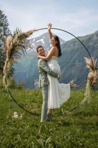 Ein glückliches Paar posiert für ein Hochzeitsfoto im Freien vor einem großen, runden Blumenbogen. Der Bräutigam in einem hellgrünen Anzug hebt die lächelnde Braut, die ein weißes Kleid trägt, hoch, während sie auf einem grasbewachsenen Hügel mit Bergen im Hintergrund stehen.