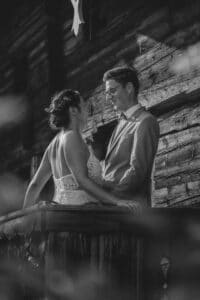 Ein Schwarzweißfoto zeigt ein Paar im Hochzeitskleid auf einem Holzbalkon, wie es sich vor einer rustikalen Kulisse liebevoll ansieht – ein bezaubernder Moment, der die Essenz einer Hochzeit wunderschön einfängt.