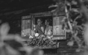 Ein Schwarzweißfoto eines lächelnden Paares, das aus einem offenen Holzfenster lehnt, umgeben von Blumen in einem Blumenkasten. Der rustikale Holzgebäudehintergrund vermittelt ein zeitloses Gefühl und fängt die Freude einer Hochzeit und die Traumkulisse für Heiraten perfekt ein.