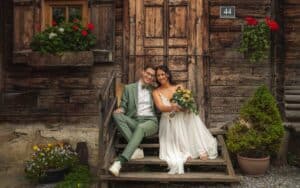 Ein Paar in Hochzeitskleidung sitzt auf Holzstufen vor einem rustikalen Gebäude und verkörpert die Essenz einer Traumhochzeit. Der Bräutigam trägt einen grünen Anzug, während die Braut einen Strauß gelber und weißer Blumen hält. Blumentöpfe und ein kleiner Baum verleihen dieser bezaubernden Szene Charme.
