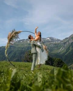 Ein Paar feiert seine Hochzeit im Freien mit Bergen im Hintergrund. Der Bräutigam hebt die Braut hoch, die ein fließendes weißes Kleid trägt, während sie lächelt und winkt. Sie stehen neben einem runden Blumenbogen auf einem grasbewachsenen Hügel, eine bezaubernde Szene, die jede Hochzeitsplanung inspirieren würde.