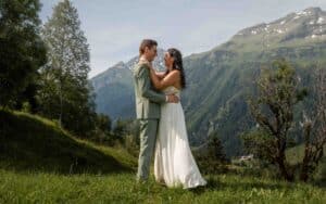 Auf einer malerischen Wiese mit Bergkulisse feiert ein Paar seine Hochzeit, umarmt sich inmitten des leuchtenden Grüns. Der Mann trägt einen grünen Anzug, während die Frau unter dem klaren Himmel ein weißes Kleid trägt und damit die Essenz ihres von Liebe erfüllten Tages einfängt.