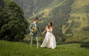 Ein Paar im Hochzeitskleid hält sich an den Händen und geht auf einem grasbedeckten Hügel spazieren, umgeben von üppigem Grün und Bergen. Der Bräutigam trägt einen grauen Anzug und die Braut ein weißes Kleid. Beide wirken fröhlich und entspannt und verkörpern die Gelassenheit ihrer Hochzeit inmitten der Pracht der Natur.