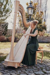 Eine Frau in einem grünen Kleid spielt Harfe im Freien in einem Garten, perfekt zum Heiraten. Sie sitzt auf einer Holzbank an einem Kopfsteinpflasterweg, umgeben von Grün und einer schwarzen Straßenlaterne, und schafft eine bezaubernde Atmosphäre für jede Hochzeit.