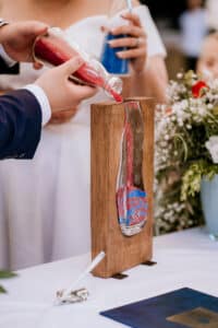 Bei ihrer Hochzeitszeremonie schüttet ein Paar farbigen Sand, rot und blau, in einen vertikalen Holzrahmen mit Glaseinsatz, was die Einheit symbolisiert. Blumen und ein Stift liegen auf dem Tisch neben dem Rahmen und verkörpern die Essenz einer durchdachten Hochzeitsplanung.